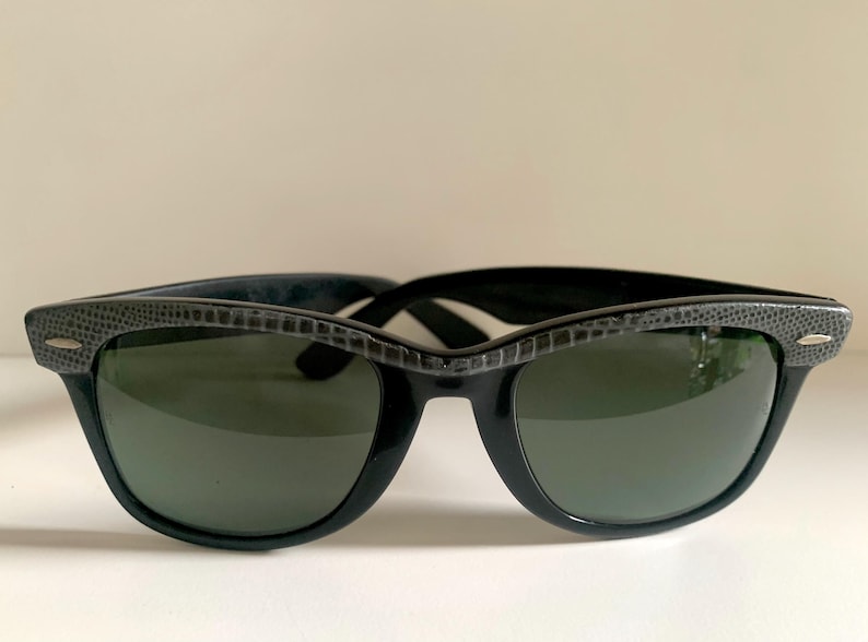 May include: Black sunglasses with a textured, dark gray upper frame and dark green lenses. The sunglasses have a classic, retro design, suitable for both men and women. The frames are made of a glossy black material.