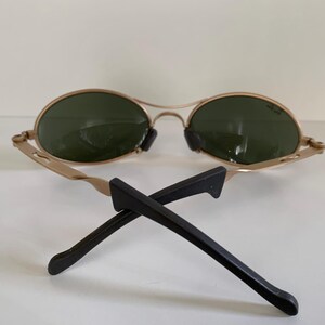 May include: Gold-framed sunglasses with oval green lenses and black temples. The sunglasses are resting on a white surface. The temples are crossed.