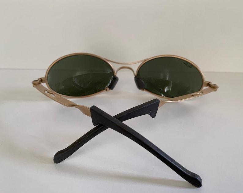 May include: Gold-framed sunglasses with oval green lenses and black temples. The sunglasses are resting on a white surface. The temples are crossed.
