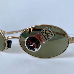 May include: Gold-tone sunglasses with oval lenses and a curved frame. The lenses are a muted green color. A sticker on the right lens features the text "SPORT" and the Olympic rings logo. The sunglasses are designed for sports and outdoor activities.