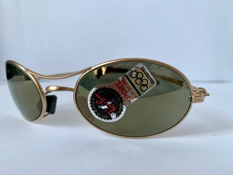 May include: Gold-tone sunglasses with oval lenses and a curved frame. The lenses are a muted green color. A sticker on the right lens features the text "SPORT" and the Olympic rings logo. The sunglasses are designed for sports and outdoor activities.