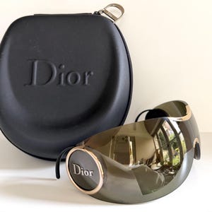 Può includere: Occhiali da sole verde oliva con dettagli dorati e il marchio "Dior". Accanto, una custodia nera con il logo "Dior" in rilievo.