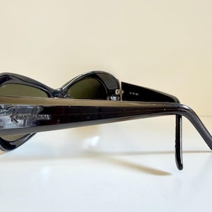 May include: Black Ray-Ban sunglasses with dark green lenses. The sunglasses have a thick, glossy black frame and the Ray-Ban logo is visible on the temple. The sunglasses are resting on a white surface.