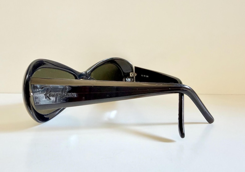 May include: Black Ray-Ban sunglasses with dark green lenses. The sunglasses have a thick, glossy black frame and the Ray-Ban logo is visible on the temple. The sunglasses are resting on a white surface.