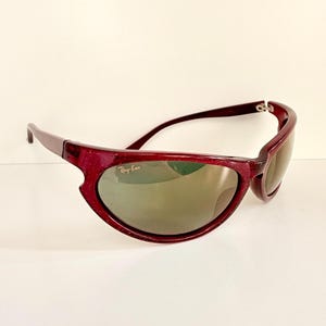 May include: Maroon-colored Ray-Ban sunglasses with a speckled finish. The sunglasses have a curved frame and tinted lenses. The Ray-Ban logo is visible on the lens. The sunglasses are set against a white background.