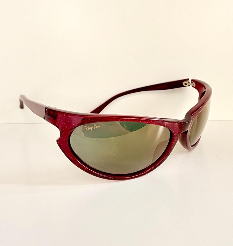 May include: Maroon-colored Ray-Ban sunglasses with a speckled finish. The sunglasses have a curved frame and tinted lenses. The Ray-Ban logo is visible on the lens. The sunglasses are set against a white background.