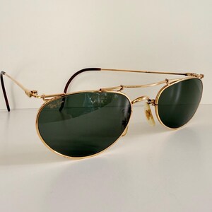 K&ouml;nnte beinhalten: Goldfarbene Sonnenbrille mit dunkelgr&uuml;nen Gl&auml;sern. Die Sonnenbrille hat einen Doppelsteg und einen klassischen Aviator-Stil. Die B&uuml;gel sind d&uuml;nn und reichen bis zu den Ohren. Das Ray-Ban-Logo ist auf dem Glas sichtbar.