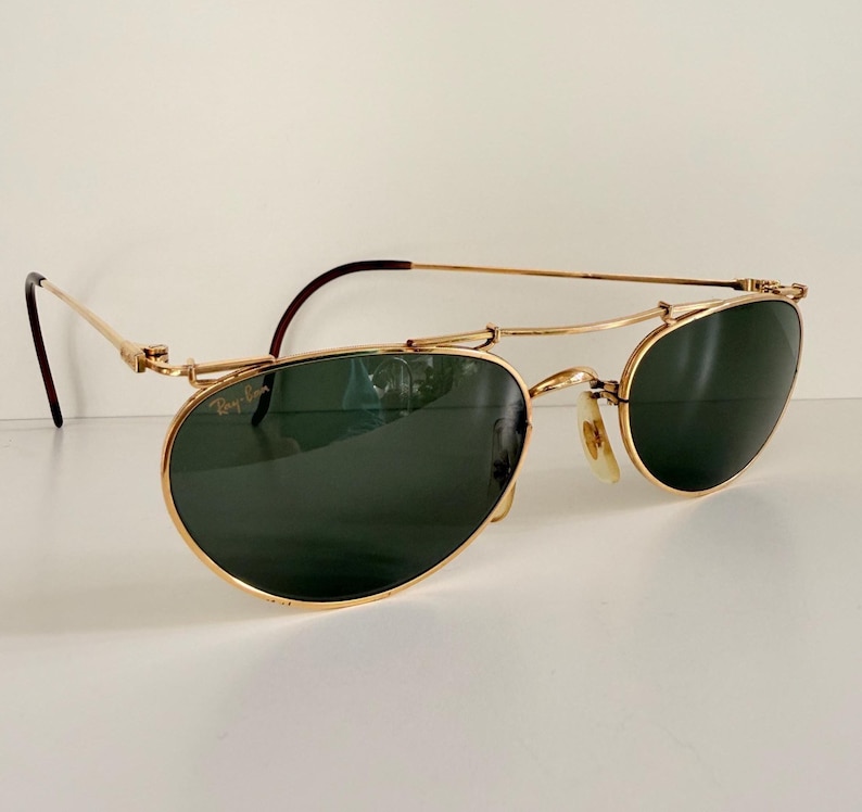 K&ouml;nnte beinhalten: Goldfarbene Sonnenbrille mit dunkelgr&uuml;nen Gl&auml;sern. Die Sonnenbrille hat einen Doppelsteg und einen klassischen Aviator-Stil. Die B&uuml;gel sind d&uuml;nn und reichen bis zu den Ohren. Das Ray-Ban-Logo ist auf dem Glas sichtbar.