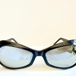 May include: Black sunglasses with a thick, curved frame and reflective lenses. The sunglasses have a retro, oversized design. The lenses reflect the surrounding environment. The sunglasses are a fashion accessory.