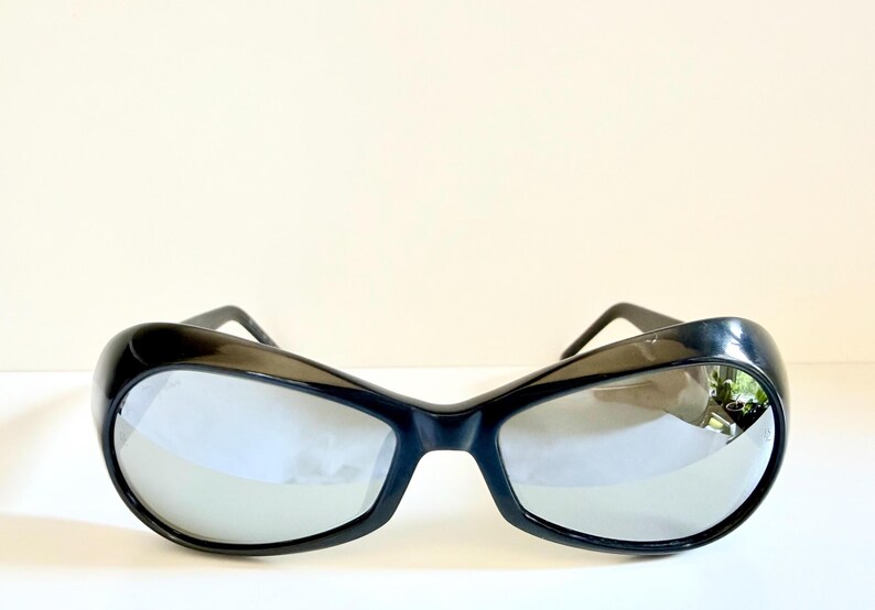 May include: Black sunglasses with a thick, curved frame and reflective lenses. The sunglasses have a retro, oversized design. The lenses reflect the surrounding environment. The sunglasses are a fashion accessory.