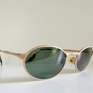 Puede incluir: Gafas de sol Ray-Ban doradas con lentes ovaladas y una barra superior sutil. Las lentes son de color verde oscuro y las patillas tienen un patr&oacute;n de carey. El logotipo de Ray-Ban es visible.