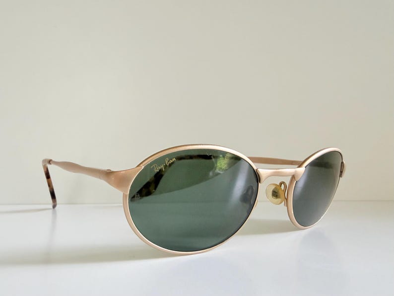 Puede incluir: Gafas de sol Ray-Ban doradas con lentes ovaladas y una barra superior sutil. Las lentes son de color verde oscuro y las patillas tienen un patr&oacute;n de carey. El logotipo de Ray-Ban es visible.