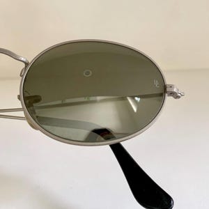 Puede incluir: Gafas de sol ovaladas plateadas con lentes verdes. Las gafas tienen una patilla de pl&aacute;stico negro.