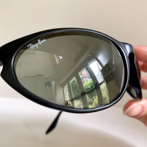 May include: Black sunglasses with a rounded, oval-shaped frame and dark tinted lenses. The upper left lens features the white Ray-Ban logo printed on the surface. The frame has a matte finish and the lenses show a reflection of a room interior.