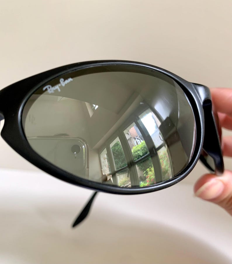 May include: Black sunglasses with a rounded, oval-shaped frame and dark tinted lenses. The upper left lens features the white Ray-Ban logo printed on the surface. The frame has a matte finish and the lenses show a reflection of a room interior.