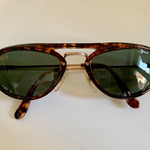 Puede incluir: Gafas de sol aviador de carey con detalles dorados y lentes verdes. Las gafas tienen el logotipo de Ray-Ban en la lente izquierda.