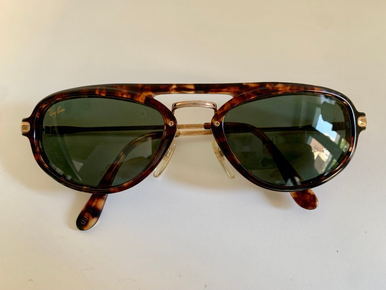 Puede incluir: Gafas de sol aviador de carey con detalles dorados y lentes verdes. Las gafas tienen el logotipo de Ray-Ban en la lente izquierda.