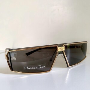 Peut inclure: Lunettes de soleil rectangulaires avec des verres fonc&eacute;s et des montures dor&eacute;es. Les lunettes de soleil portent un logo Christian Dior dor&eacute; dans le coin sup&eacute;rieur gauche. Le texte "Christian Dior" est imprim&eacute; sur un autocollant blanc sur le verre gauche.