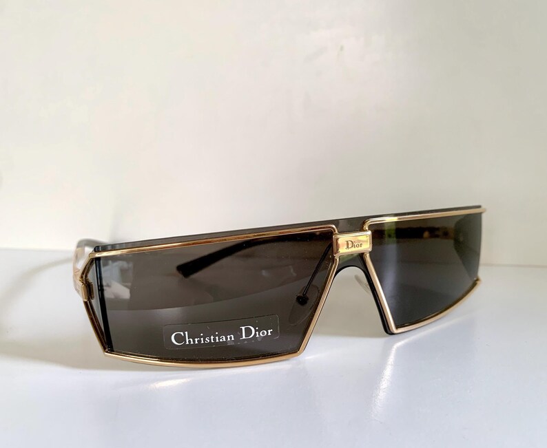 Peut inclure: Lunettes de soleil rectangulaires avec des verres fonc&eacute;s et des montures dor&eacute;es. Les lunettes de soleil portent un logo Christian Dior dor&eacute; dans le coin sup&eacute;rieur gauche. Le texte "Christian Dior" est imprim&eacute; sur un autocollant blanc sur le verre gauche.