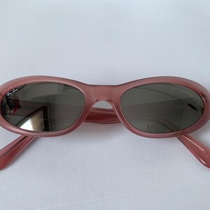 Peut inclure: Lunettes de soleil ovales de couleur rose translucide. Les verres sont gris fonc&eacute; et les montures ont un &eacute;clat subtil. Le logo Ray-Ban est visible sur le verre gauche. Un accessoire &eacute;l&eacute;gant pour la protection des yeux.