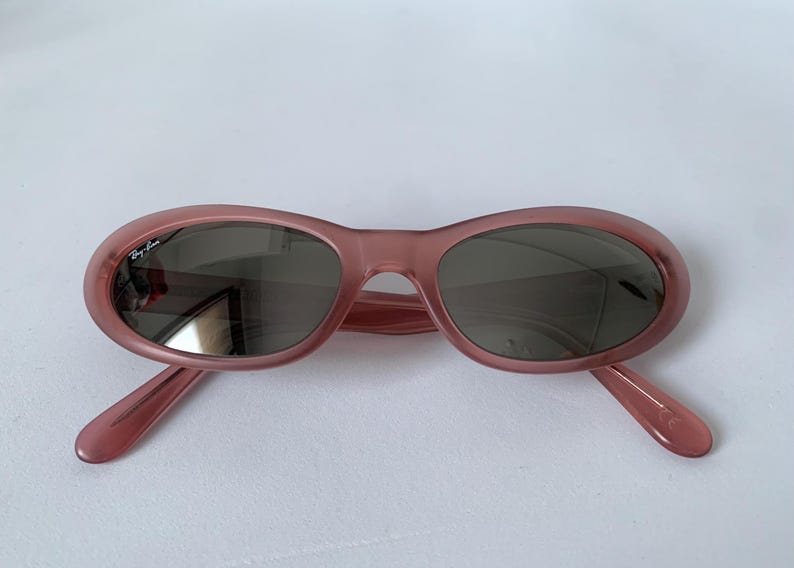 Peut inclure: Lunettes de soleil ovales de couleur rose translucide. Les verres sont gris fonc&eacute; et les montures ont un &eacute;clat subtil. Le logo Ray-Ban est visible sur le verre gauche. Un accessoire &eacute;l&eacute;gant pour la protection des yeux.