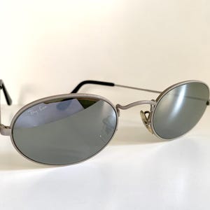 Puede incluir: Gafas de sol ovaladas de color plateado con lentes espejados. Las gafas tienen el logotipo de Ray-Ban en la lente izquierda.
