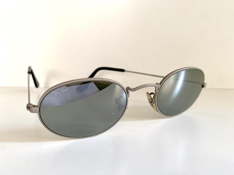Puede incluir: Gafas de sol ovaladas de color plateado con lentes espejados. Las gafas tienen el logotipo de Ray-Ban en la lente izquierda.