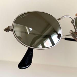 Puede incluir: Gafas de sol redondas Ray-Ban plateadas con lentes verdes. Las gafas tienen una patilla de pl&aacute;stico negro con el texto "Ray-Ban Made in Italy" impreso en ella.