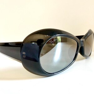 May include: Black oval sunglasses with grey lenses. The sunglasses have a thick, glossy black frame and the Ray-Ban logo is visible on the lens. The design is retro and stylish.