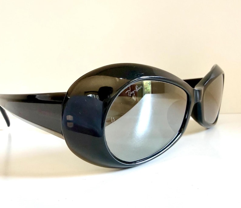 May include: Black oval sunglasses with grey lenses. The sunglasses have a thick, glossy black frame and the Ray-Ban logo is visible on the lens. The design is retro and stylish.