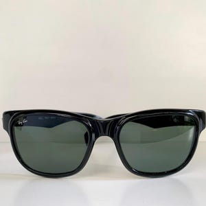 Puede incluir: Gafas de sol negras con lentes verde oscuro. Las gafas tienen una montura negra gruesa y brillante y un diseño curvo. El logotipo de Ray-Ban es visible en la lente izquierda. Las gafas están sobre un fondo blanco.