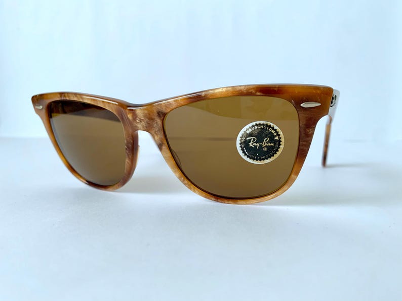 Puede incluir: Gafas de sol Ray-Ban de carey marr&oacute;n con lentes marrones. Las gafas tienen una barra de metal en el puente y una pegatina del logotipo de Ray-Ban en la lente derecha.