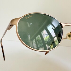 Puede incluir: Primer plano de gafas de sol Ray-Ban con montura dorada y lentes verde oscuro. El logotipo de Ray-Ban es visible en la parte superior de la lente izquierda. Las patillas son finas y tienen un patr&oacute;n de carey en los extremos.