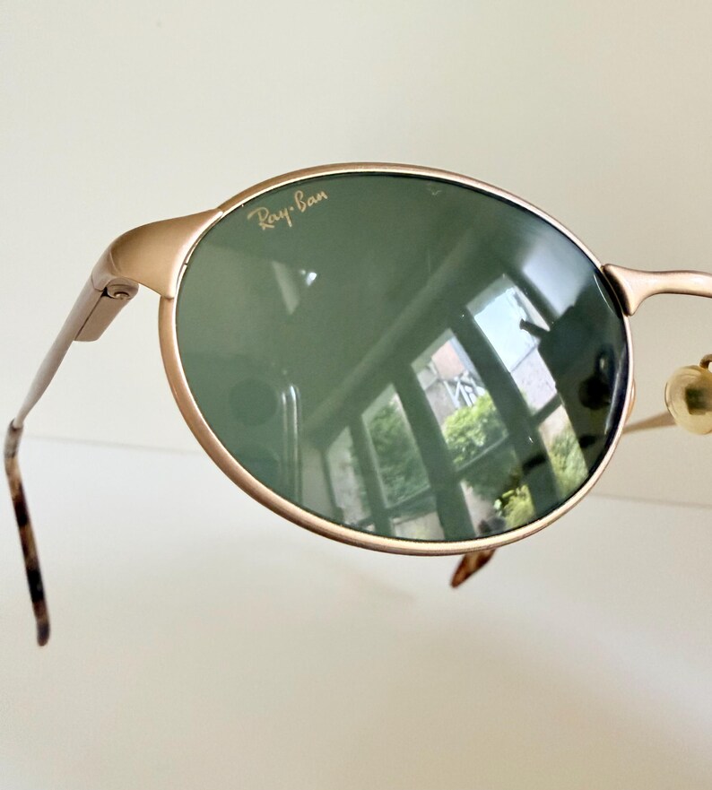 Puede incluir: Primer plano de gafas de sol Ray-Ban con montura dorada y lentes verde oscuro. El logotipo de Ray-Ban es visible en la parte superior de la lente izquierda. Las patillas son finas y tienen un patr&oacute;n de carey en los extremos.