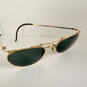 K&ouml;nnte beinhalten: Goldfarbene Sonnenbrille mit dunkelgr&uuml;nen Gl&auml;sern. Die Sonnenbrille hat ein klassisches Design mit einem d&uuml;nnen Goldrahmen und braunen B&uuml;geln. Die Gl&auml;ser sind oval und leicht gebogen. Auf wei&szlig;em Hintergrund.