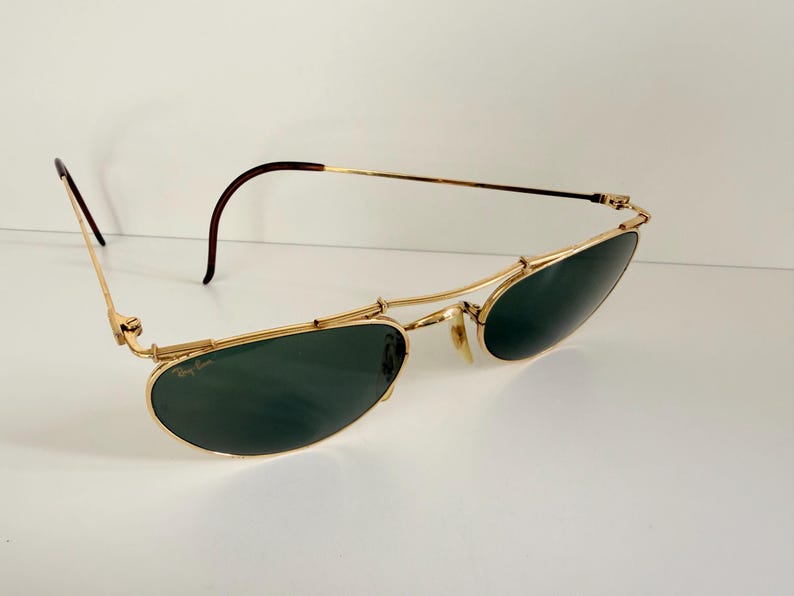 K&ouml;nnte beinhalten: Goldfarbene Sonnenbrille mit dunkelgr&uuml;nen Gl&auml;sern. Die Sonnenbrille hat ein klassisches Design mit einem d&uuml;nnen Goldrahmen und braunen B&uuml;geln. Die Gl&auml;ser sind oval und leicht gebogen. Auf wei&szlig;em Hintergrund.