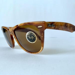 Puede incluir: Gafas de sol Ray-Ban de carey marr&oacute;n con lentes marrones. Las gafas tienen un marco de estilo Wayfarer cl&aacute;sico y un logotipo Ray-Ban dorado en la patilla derecha.