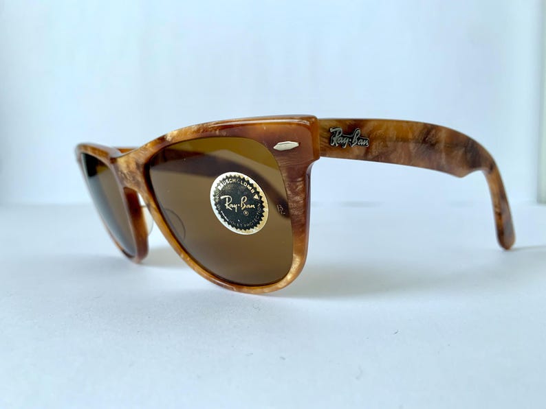 Puede incluir: Gafas de sol Ray-Ban de carey marr&oacute;n con lentes marrones. Las gafas tienen un marco de estilo Wayfarer cl&aacute;sico y un logotipo Ray-Ban dorado en la patilla derecha.