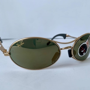 May include: Gold-framed sunglasses with oval lenses and a green tint. The Ray-Ban logo is visible on the left lens. The sunglasses have black accents on the temples and nose bridge. A sticker is on the right lens.