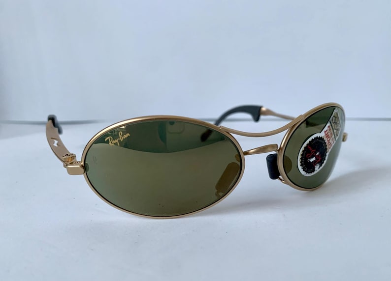 May include: Gold-framed sunglasses with oval lenses and a green tint. The Ray-Ban logo is visible on the left lens. The sunglasses have black accents on the temples and nose bridge. A sticker is on the right lens.