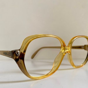 Könnte beinhalten: Eine Brille aus bernsteinfarbenem Kunststoff mit einem quadratischen Gestell. Die Brille hat einen silbernen Akzent an der Seite.