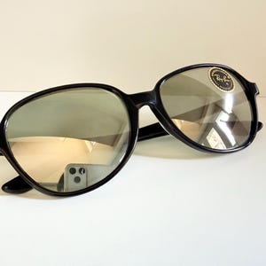 May include: Black sunglasses with a rounded frame and reflective lenses. The sunglasses have a small, circular sticker on the lens. The frames are thick and the lenses are a light gray color.
