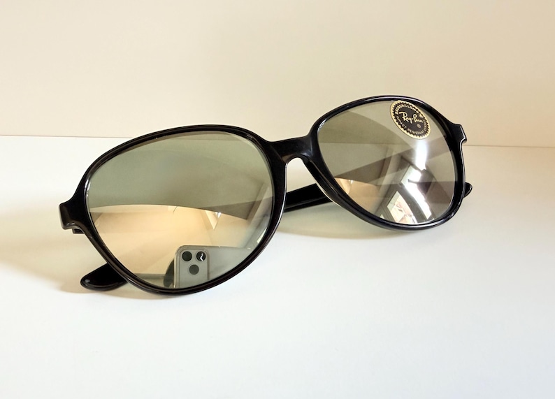 May include: Black sunglasses with a rounded frame and reflective lenses. The sunglasses have a small, circular sticker on the lens. The frames are thick and the lenses are a light gray color.