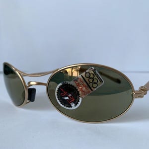 May include: Gold-framed sunglasses with oval lenses and a black nose bridge. The lenses are a muted green color. A sticker with the Olympic rings is affixed to the right lens. The sunglasses are angled, showing the side view.