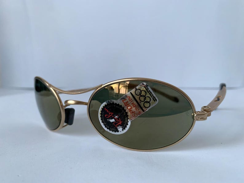 May include: Gold-framed sunglasses with oval lenses and a black nose bridge. The lenses are a muted green color. A sticker with the Olympic rings is affixed to the right lens. The sunglasses are angled, showing the side view.