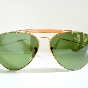 May include: Aviator-style sunglasses with a gold-colored frame and green lenses. The sunglasses have a light-colored bridge and the Ray-Ban logo is visible on the lens. The lenses are reflective.