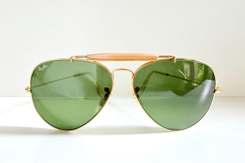 May include: Aviator-style sunglasses with a gold-colored frame and green lenses. The sunglasses have a light-colored bridge and the Ray-Ban logo is visible on the lens. The lenses are reflective.