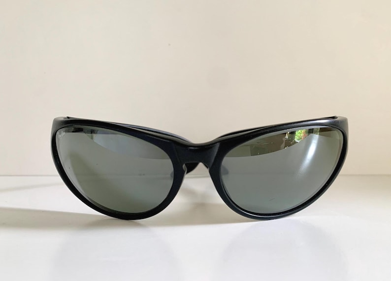 May include: Black sunglasses featuring a wraparound frame design with dark, reflective lenses. The frames are constructed from a smooth, solid black material that curves around the face, providing a wide field of vision and a sleek, continuous look.