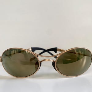 May include: Oval-shaped sunglasses with a gold-colored frame and dark lenses. The bridge and temples are black. The sunglasses are set against a white background.