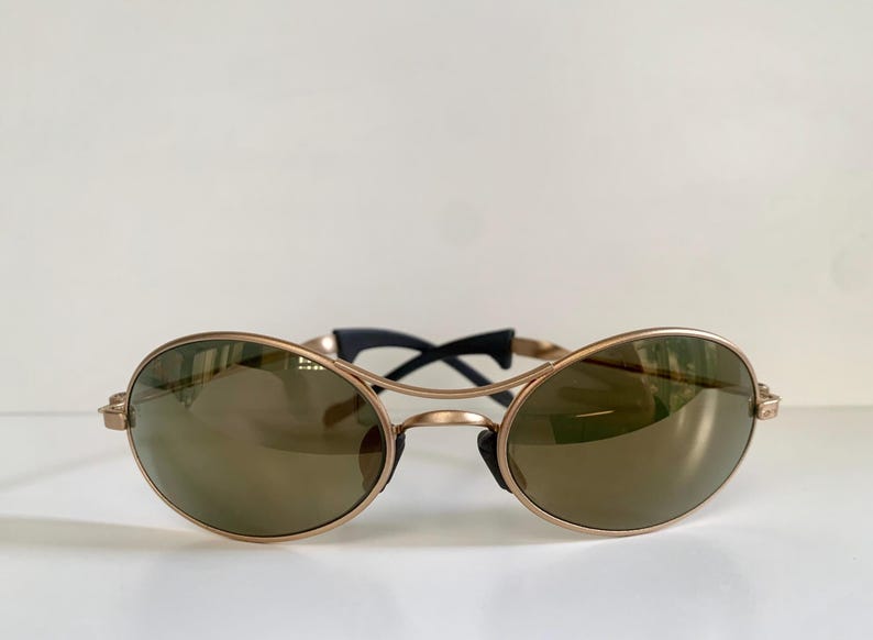 May include: Oval-shaped sunglasses with a gold-colored frame and dark lenses. The bridge and temples are black. The sunglasses are set against a white background.
